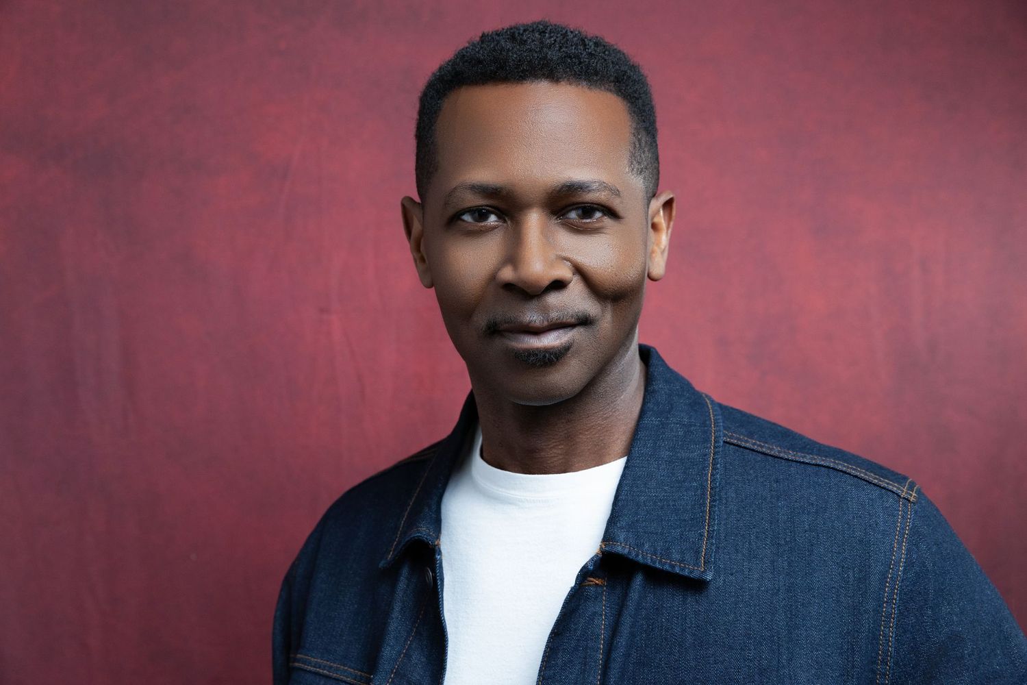 Professional headshot of person wearing denim jacket and white t-shirt against red backdrop.