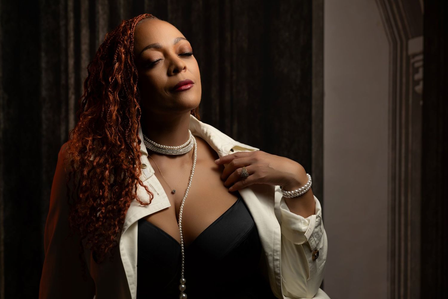 A person with long red curly hair poses in a white blazer and black top against a dark background with dramatic lighting.