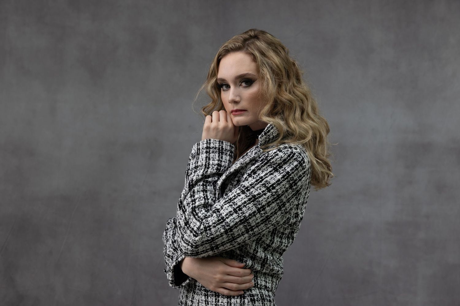 Woman in plaid blazer with wavy blonde hair poses confidently against gray studio background.