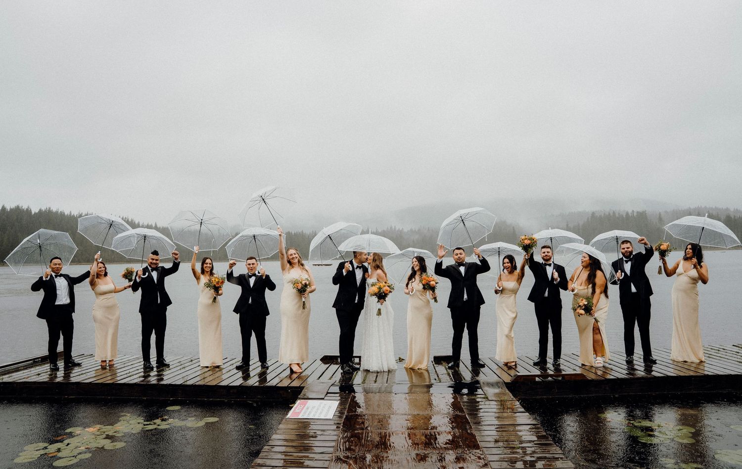Wedding party stands in a row holding white umbrellas on a rainy day by a lake with mountains in the background.