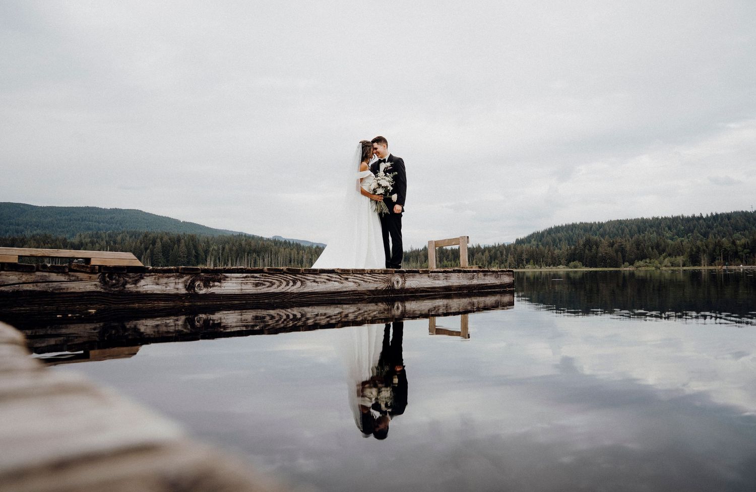 A serene lake reflects the cloudy sky with wooden docks extending into calm waters surrounded by forested hills.