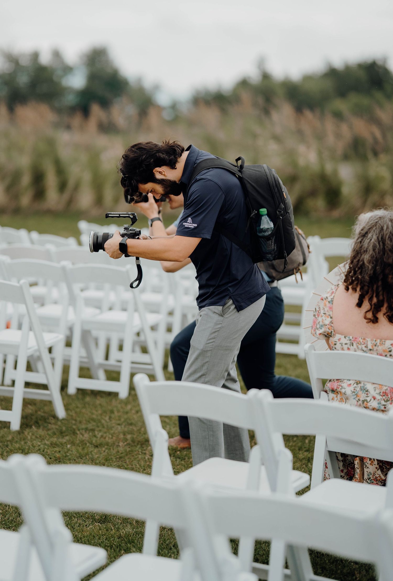 Wedding photographer crouches between white chairs to capture ceremony shots from a unique angle at outdoor venue.