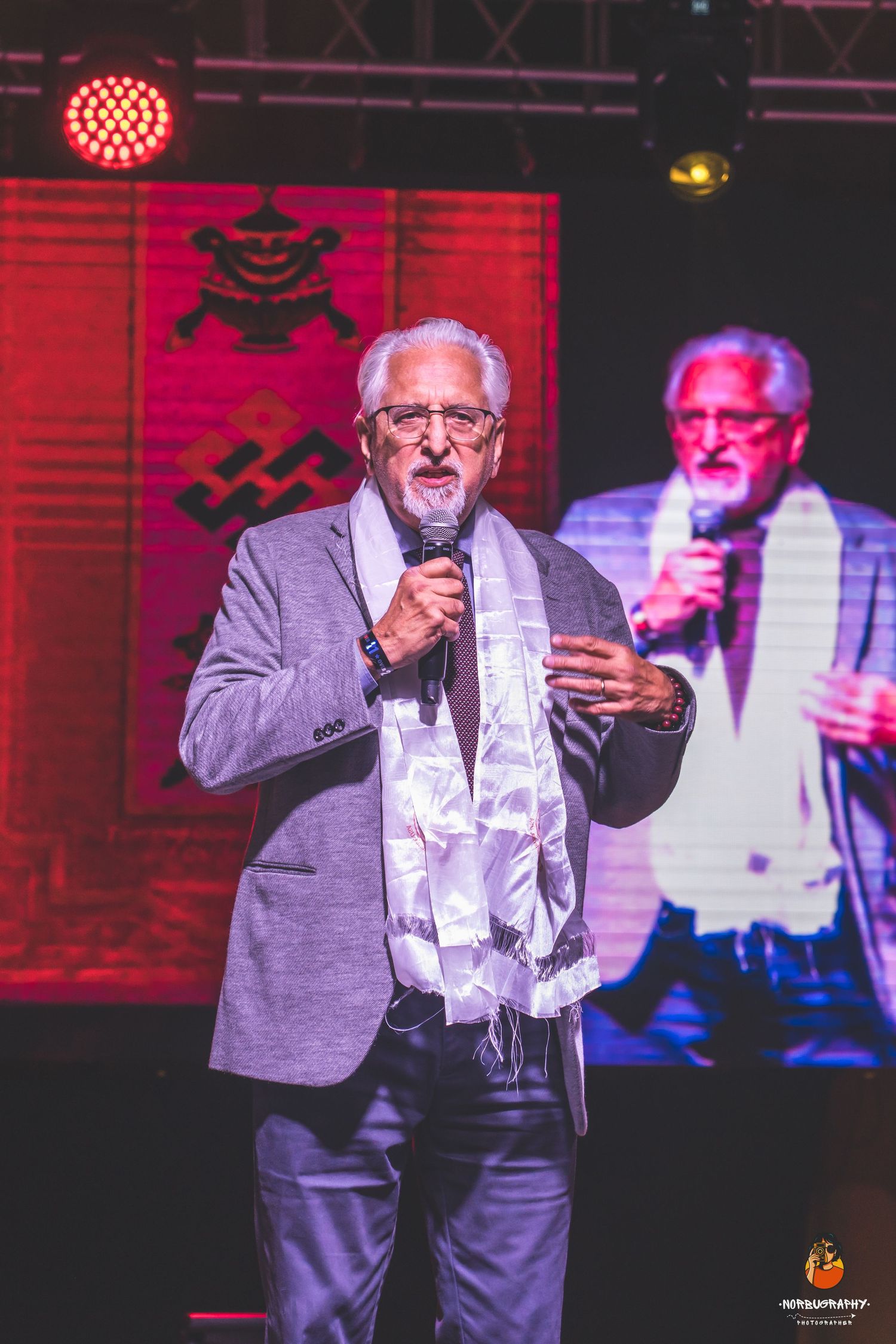 Speaker in grey suit with white scarf performs on stage with red lighting and traditional decorations.