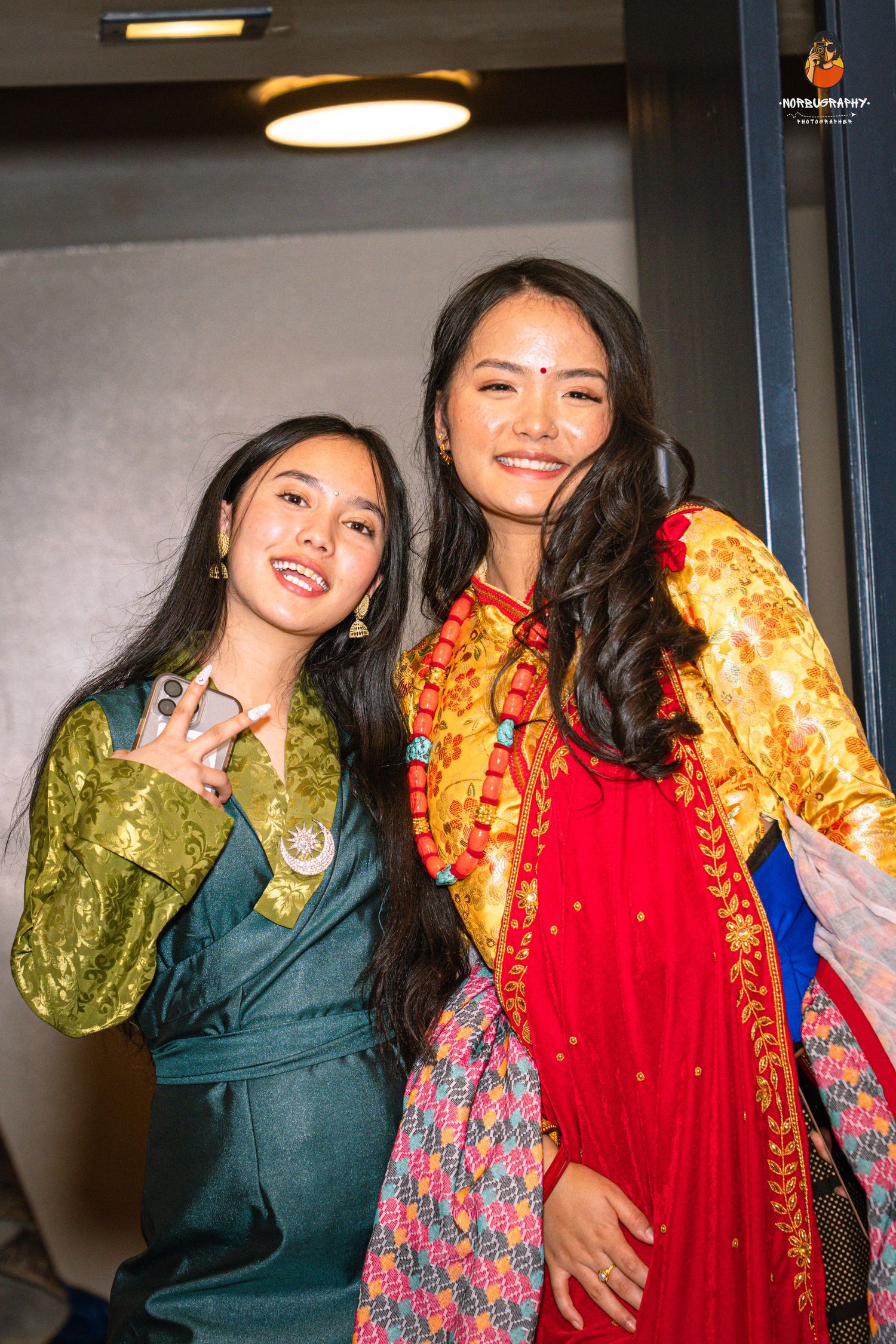 Two women in colorful traditional Asian outfits smile and pose together in an indoor setting with warm lighting.
