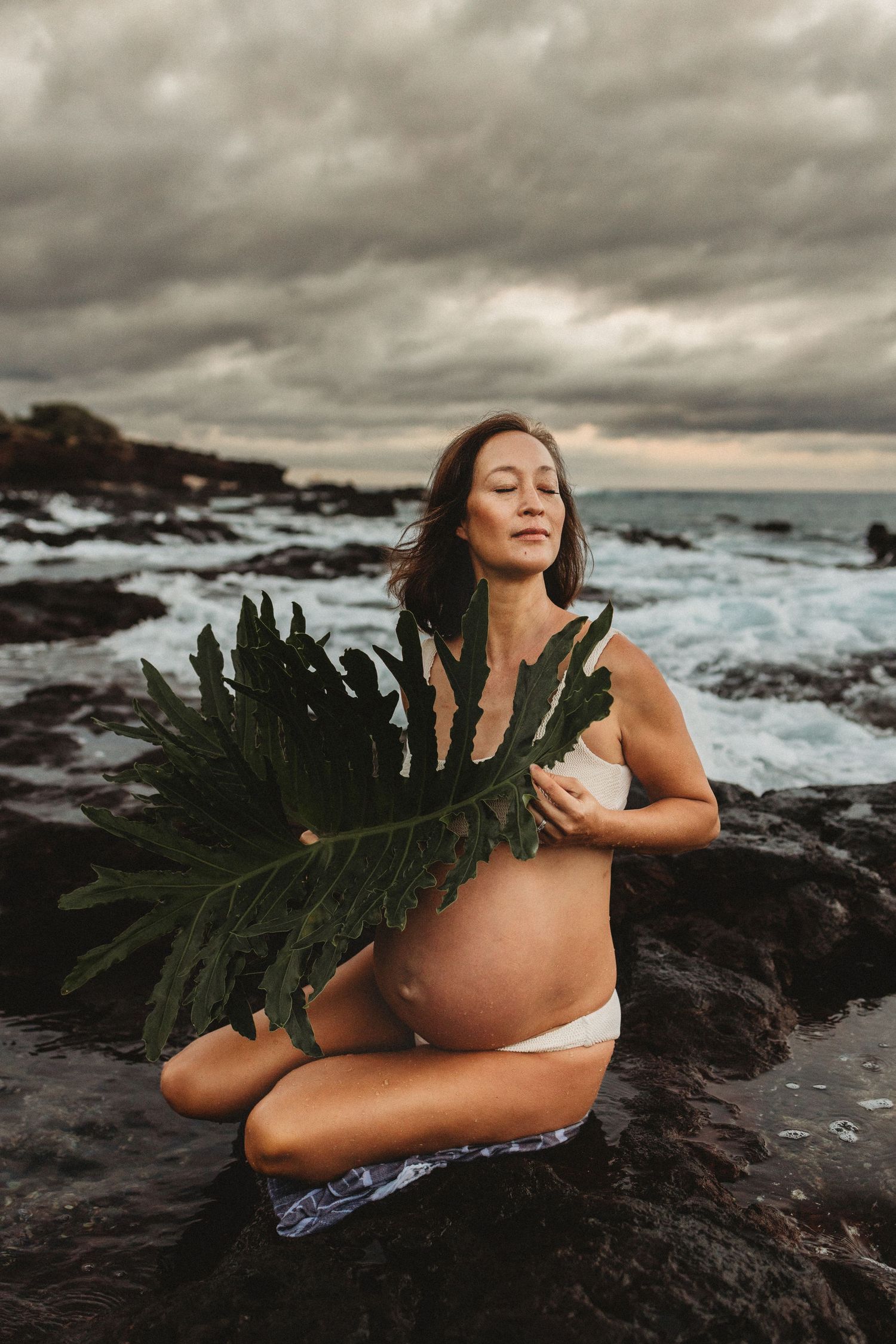 Maui maternity photography of pregnant woman sitting on a rock holding a tropical leaf.