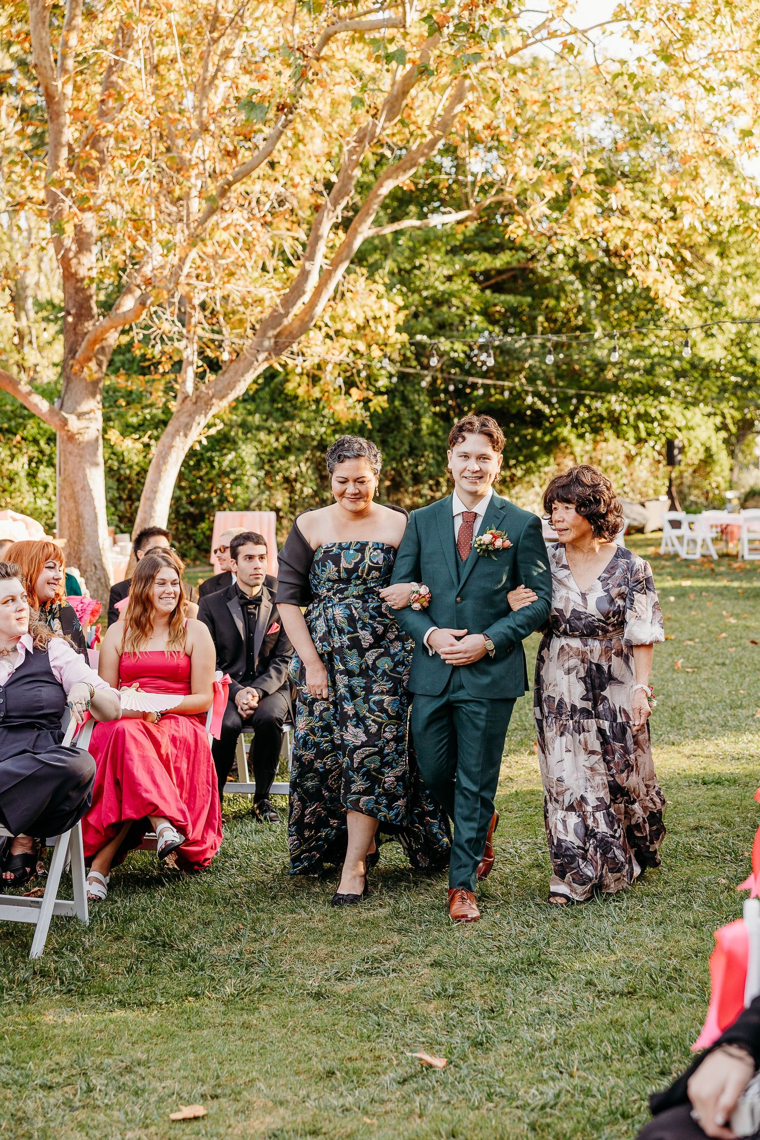 Wedding guests seated on lawn under autumn trees during outdoor ceremony.