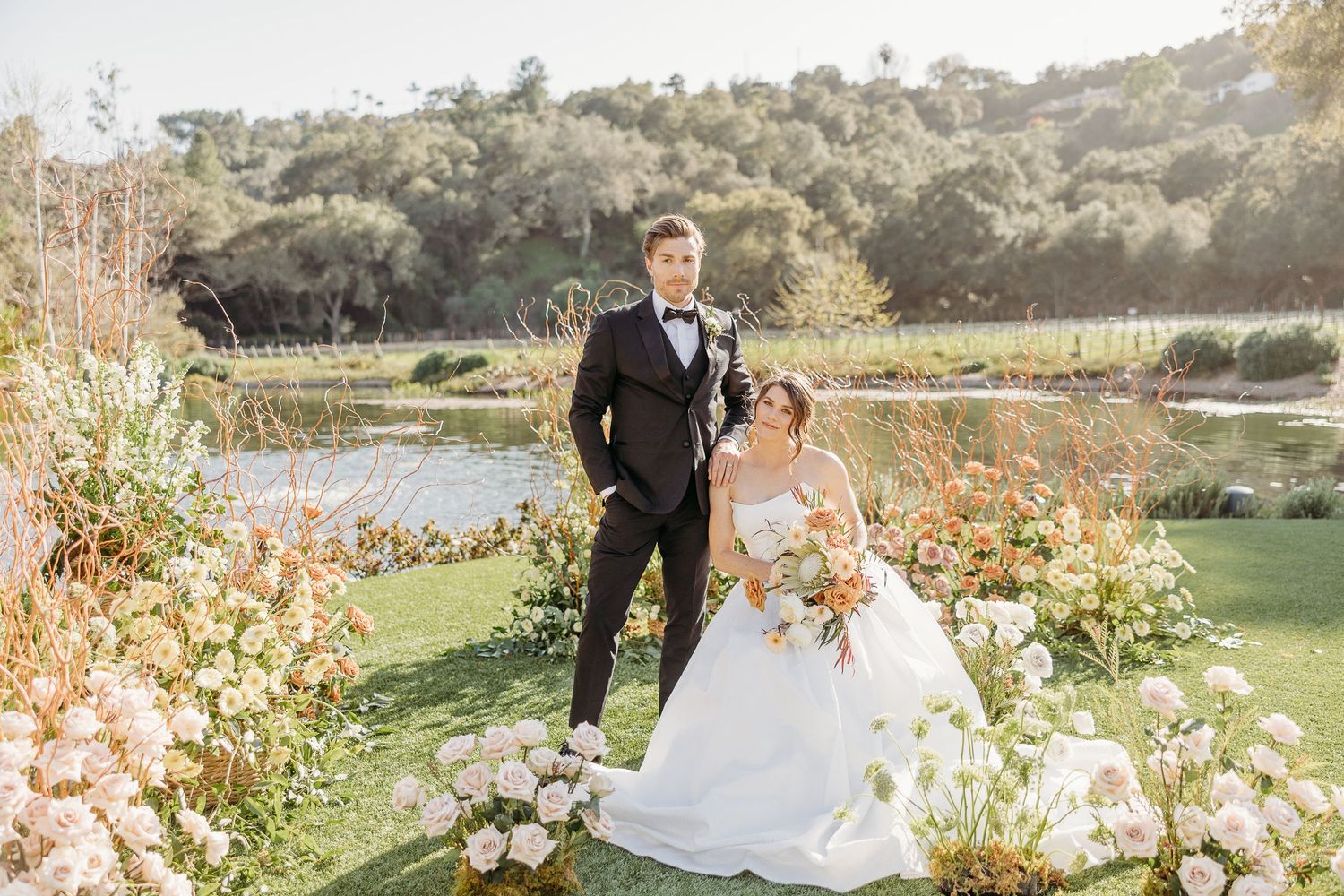 A couple poses together in a scenic garden setting surrounded by white flowers and natural greenery during golden hour.