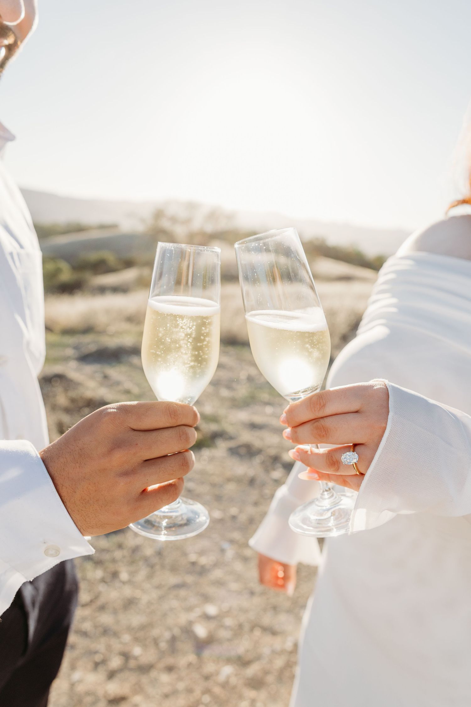 Two people holding wine glasses with white wine clink together in an outdoor setting at sunset.