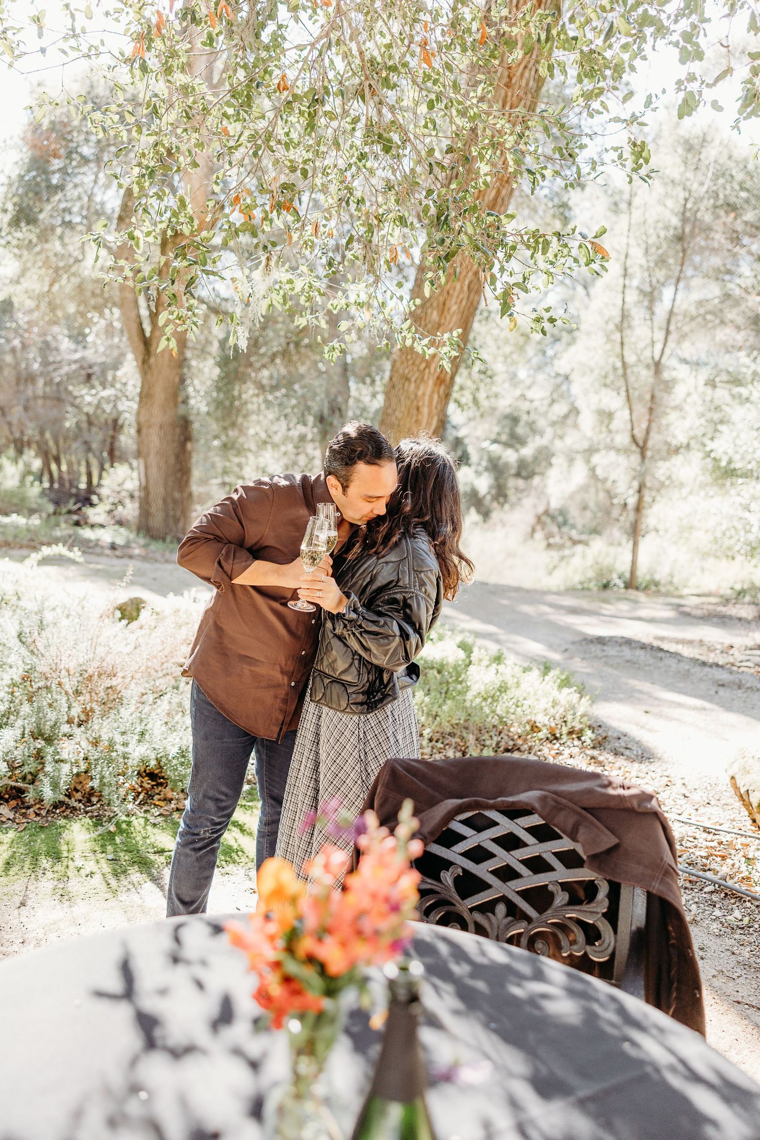 A romantic outdoor scene with a couple embracing near a floral arrangement on a table under tall trees.