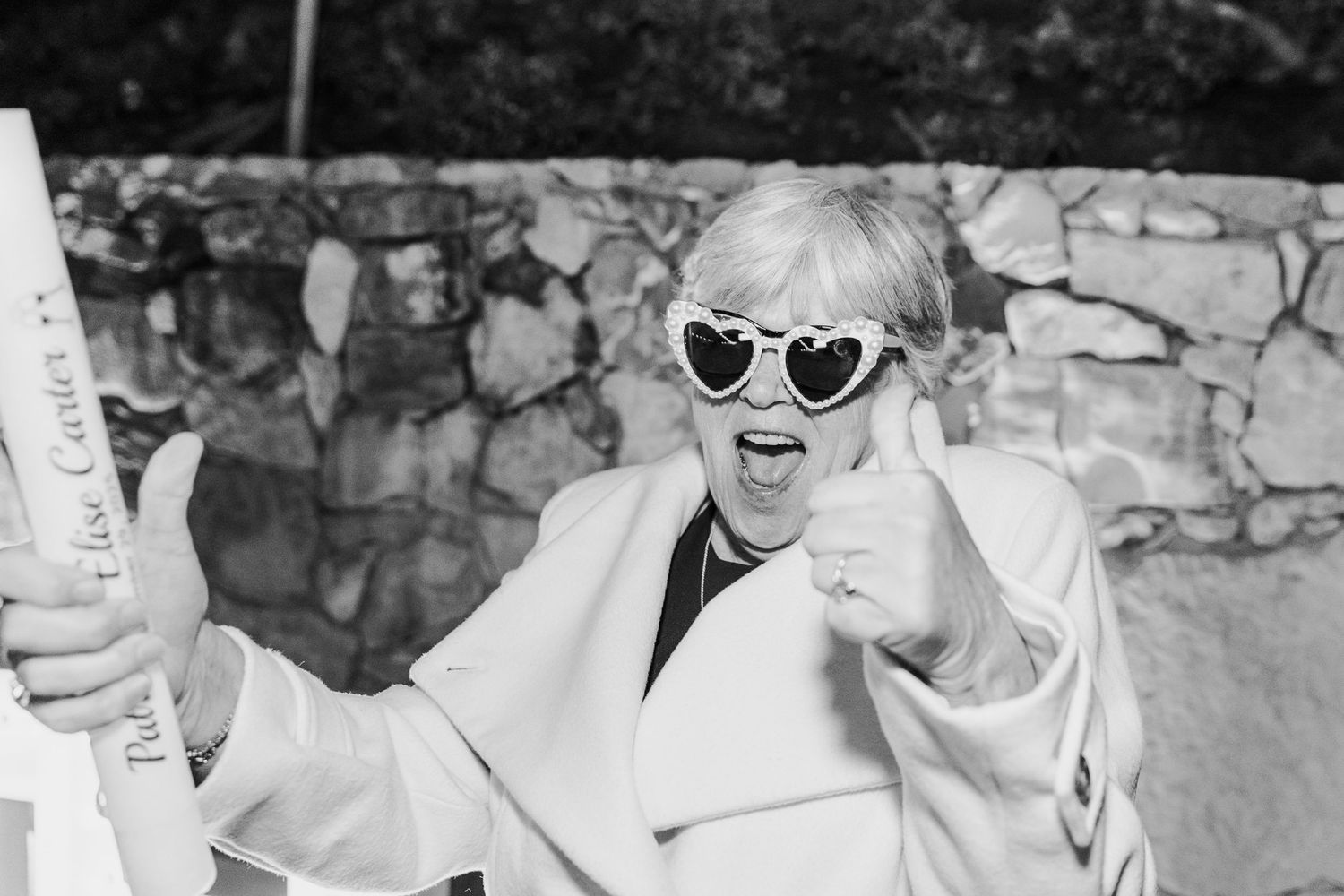 A candid black and white photo of someone in sunglasses and a white coat expressing joy.