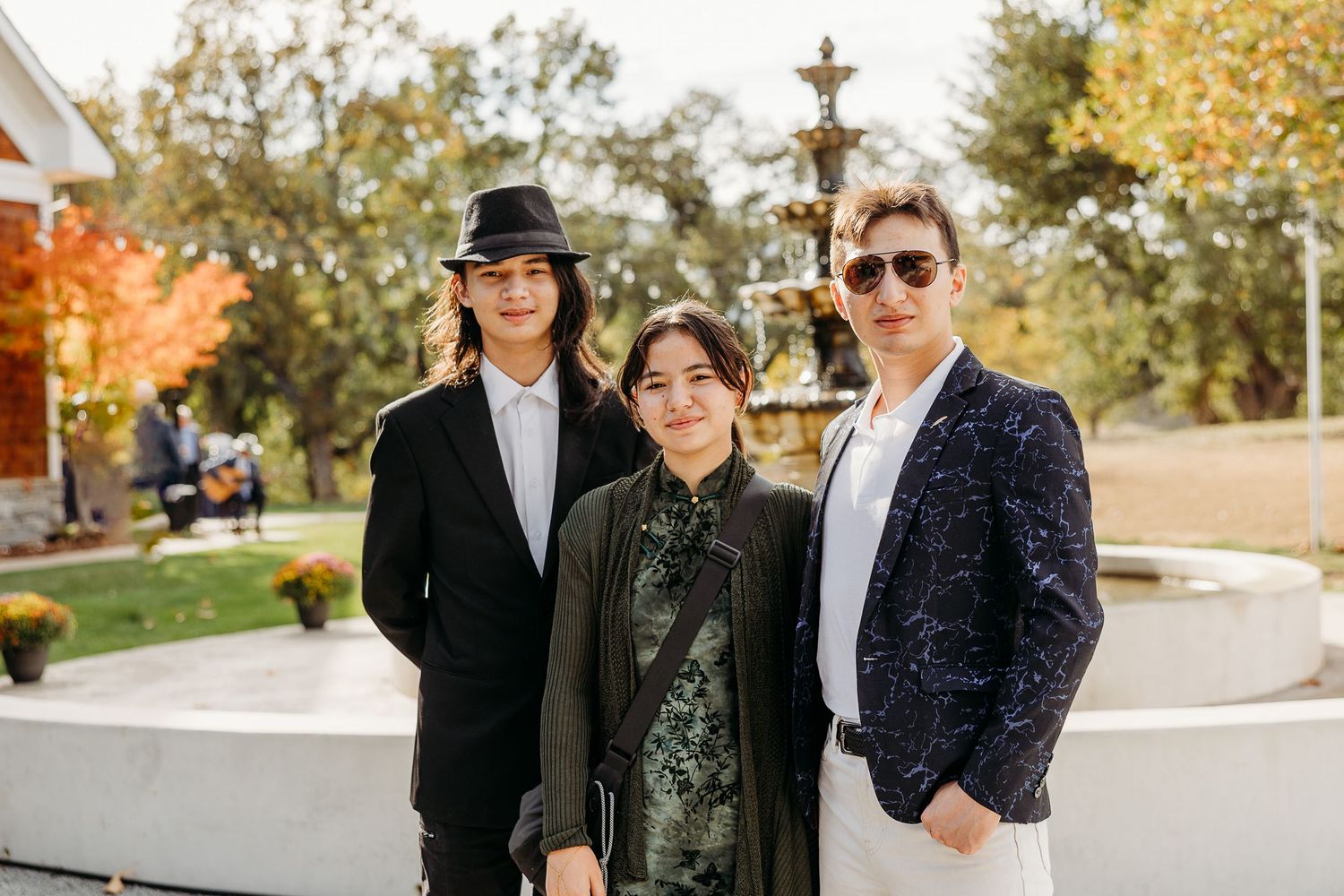 Three guests in formal attire pose outdoors at an autumn wedding.