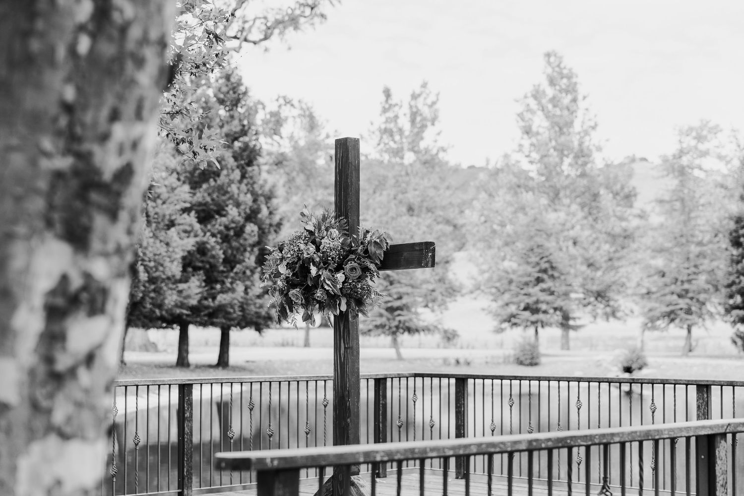 A wooden cross adorned with flowers stands on a fenced deck with trees in the misty background.