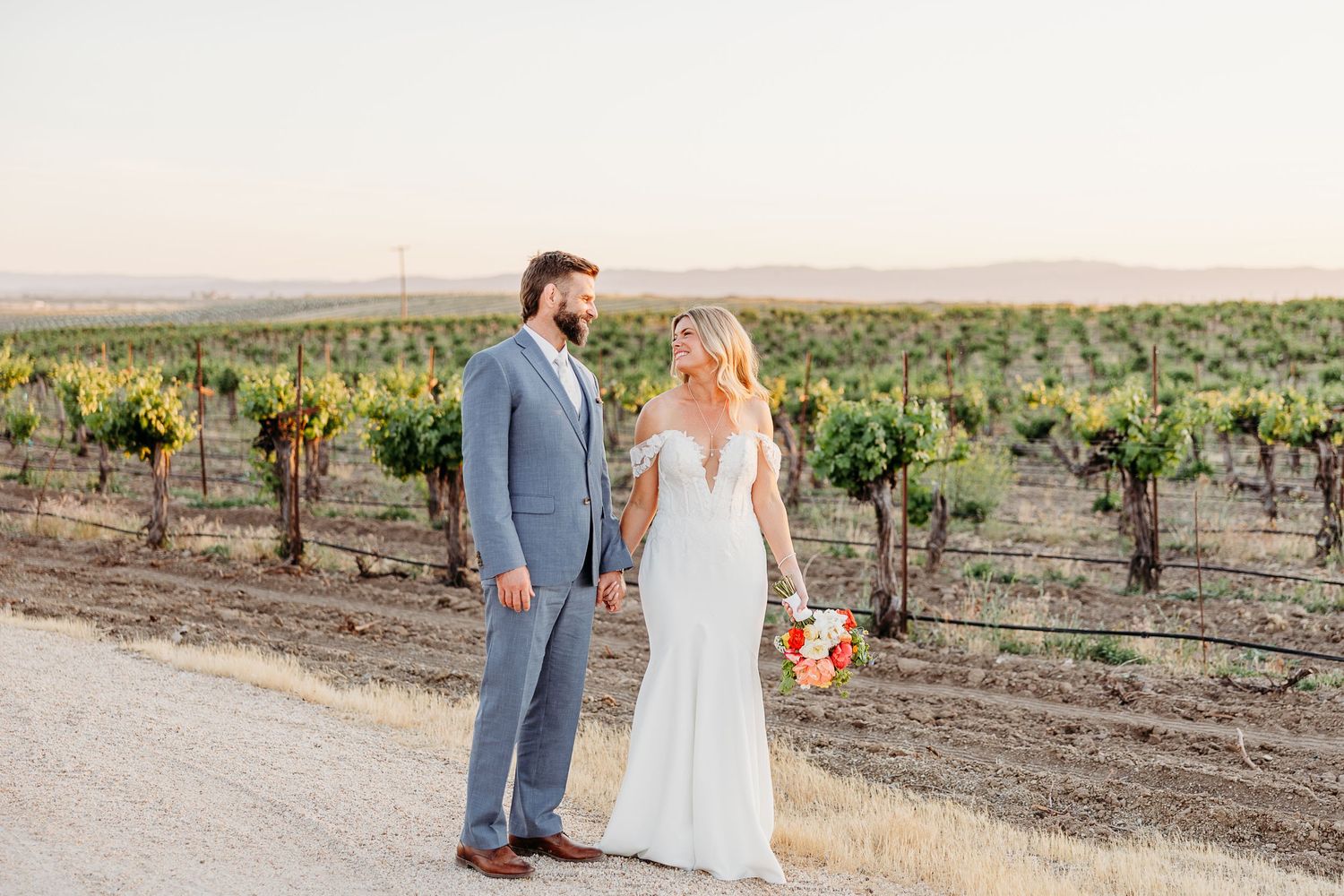 Couple in wedding attire walking through scenic vineyard rows during sunset in wine country.