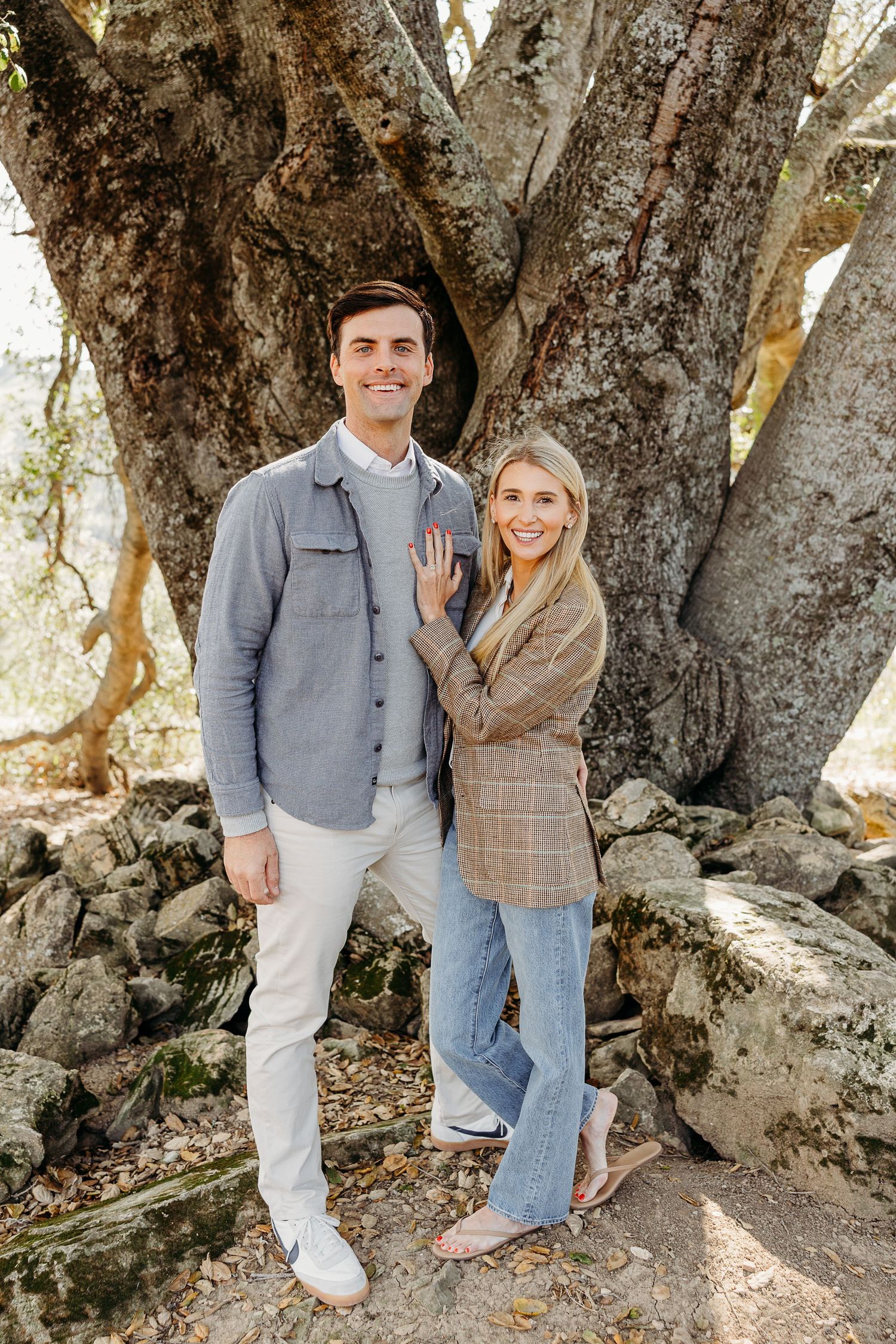 Casual couple posing together against a large tree trunk in nature.