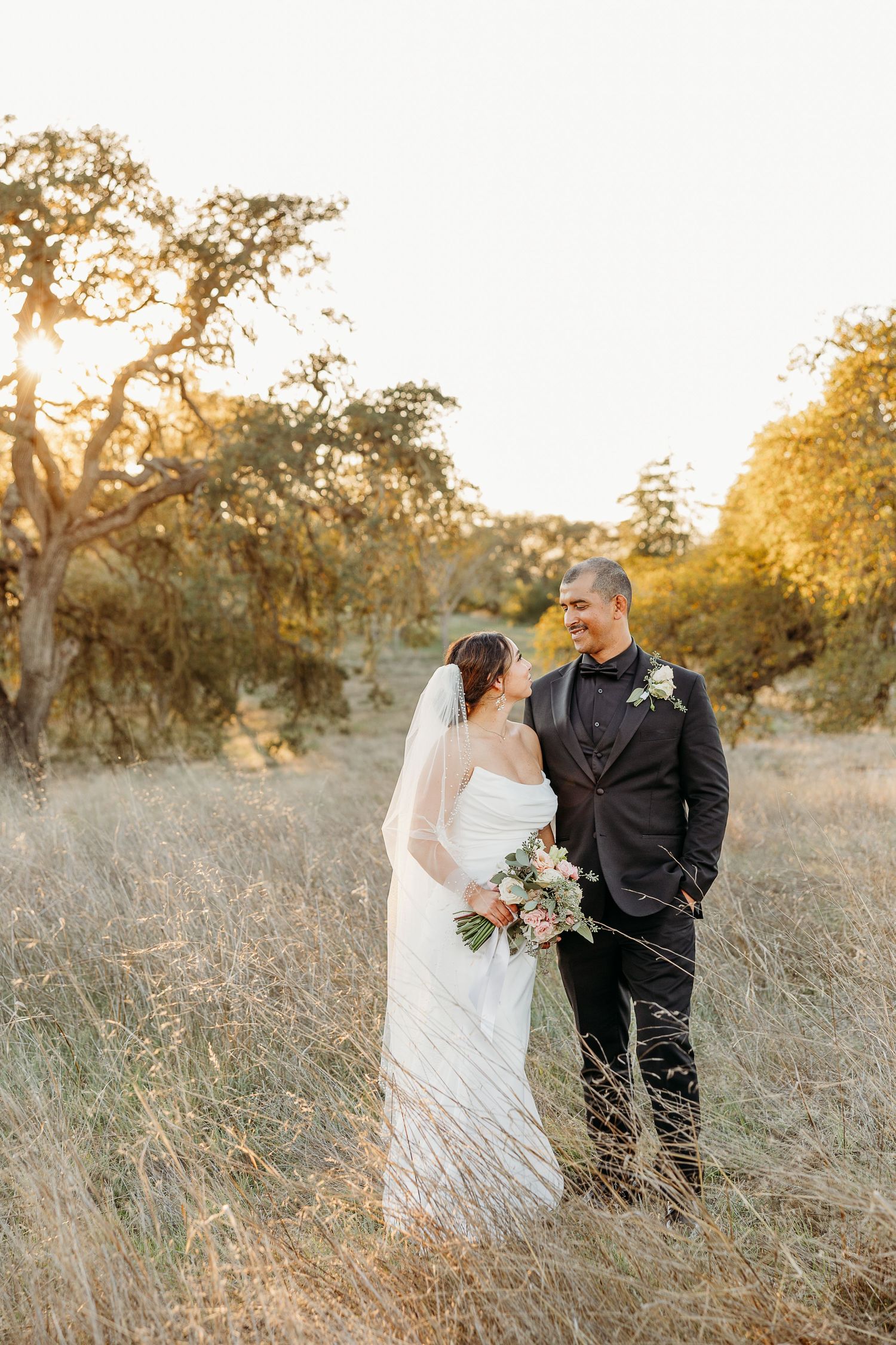 Newlywed couple poses in golden field at sunset wearing traditional wedding attire.