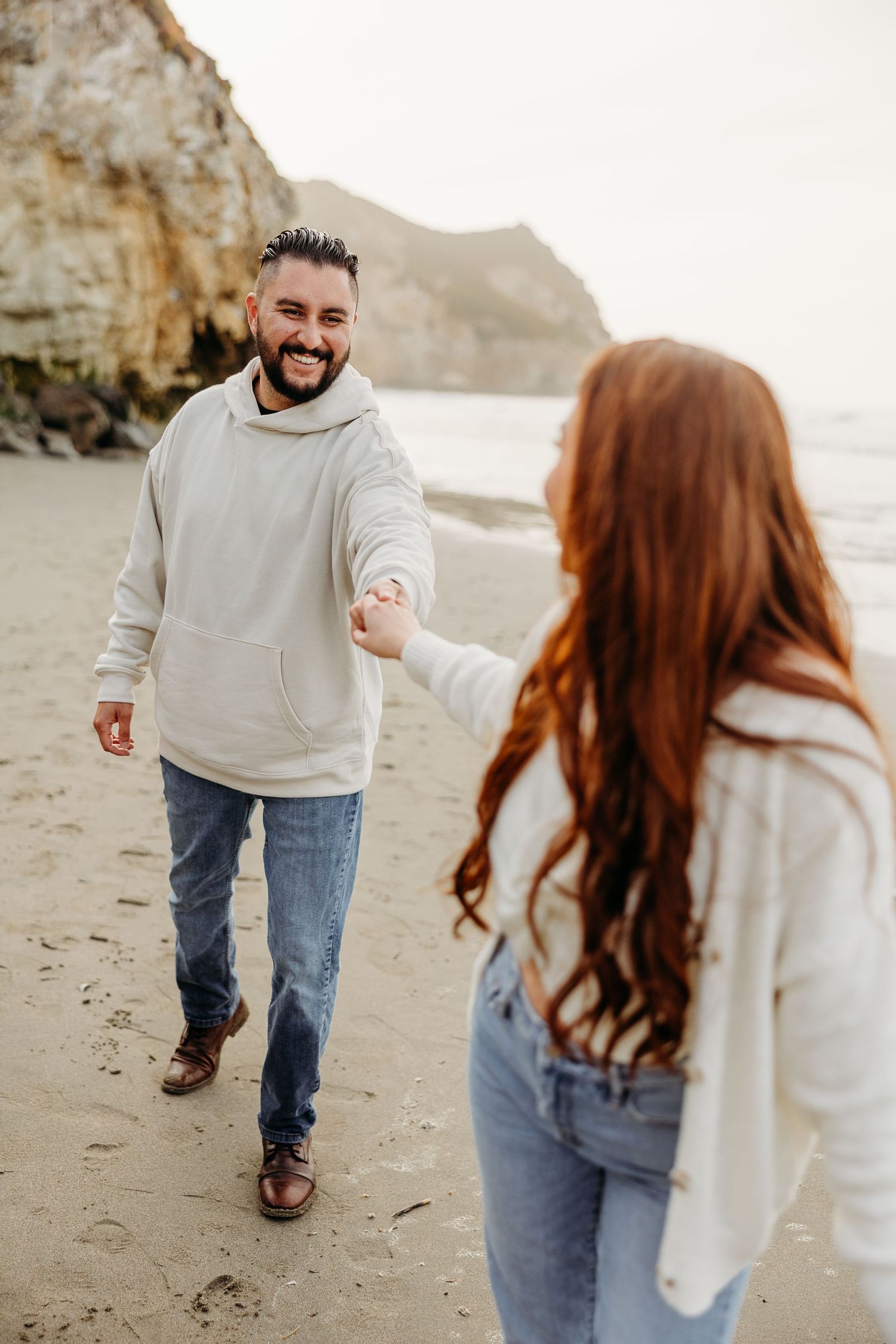 A couple wearing matching light sweaters and jeans walks together on a scenic beach with cliffs in the background.