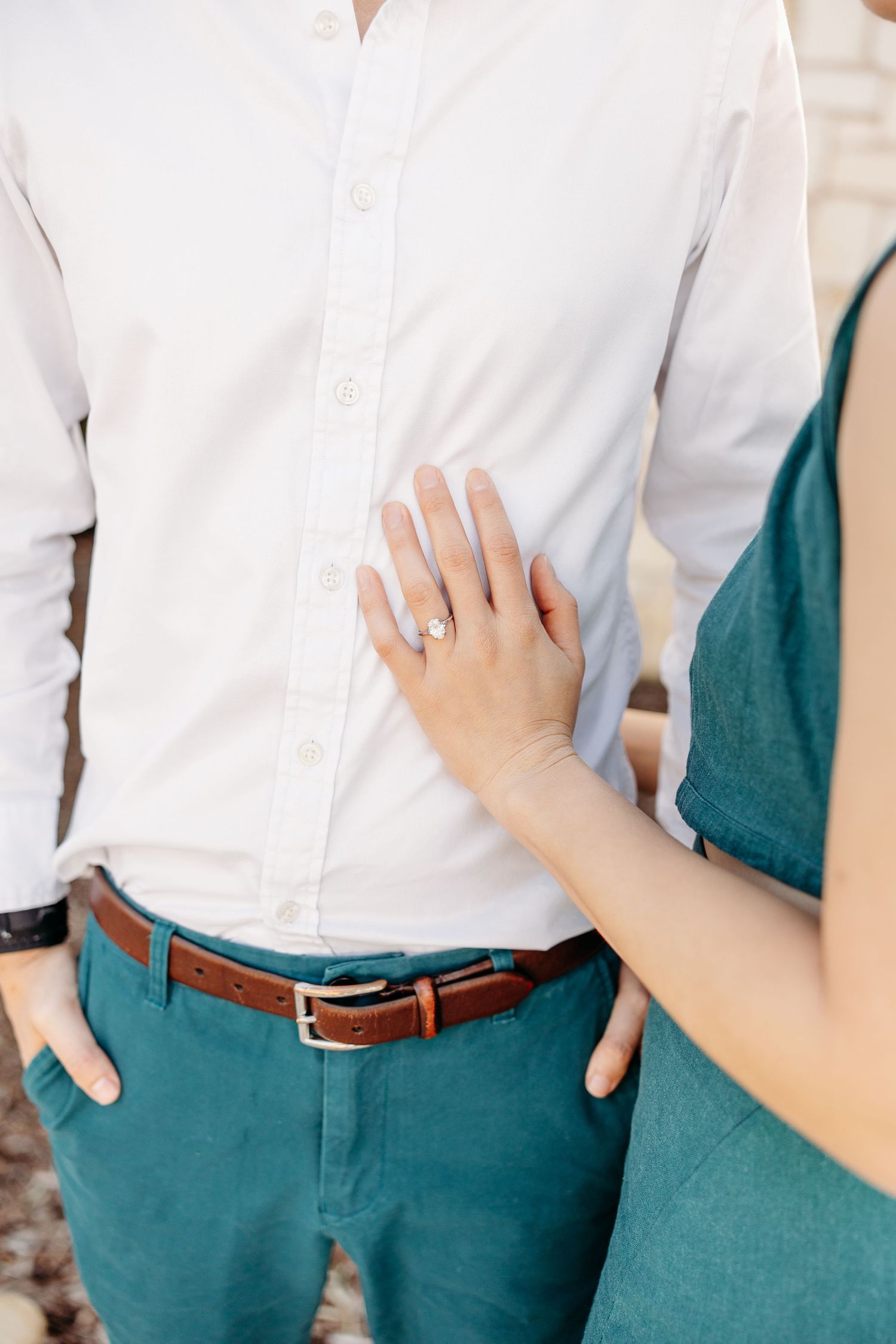 Close up view of a hand with an engagement ring resting on a white dress shirt with teal pants.