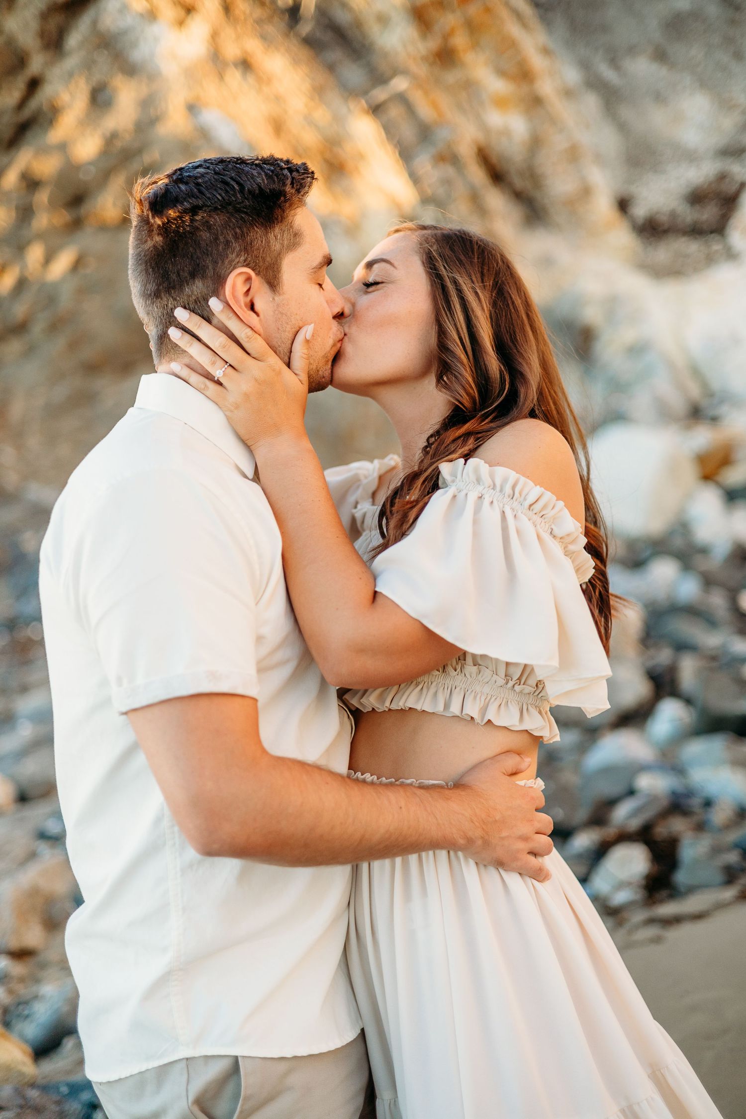 Intimate couple sharing a kiss on sandy beach with dramatic cliffs in background at sunset.