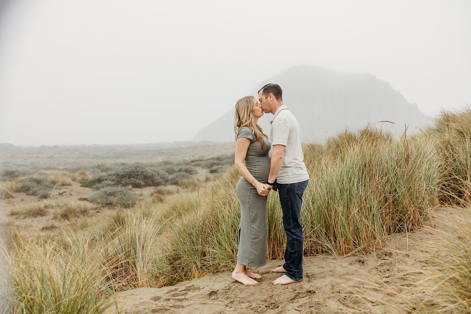 A couple embraces on sandy dunes surrounded by tall beach grass during a maternity photoshoot.