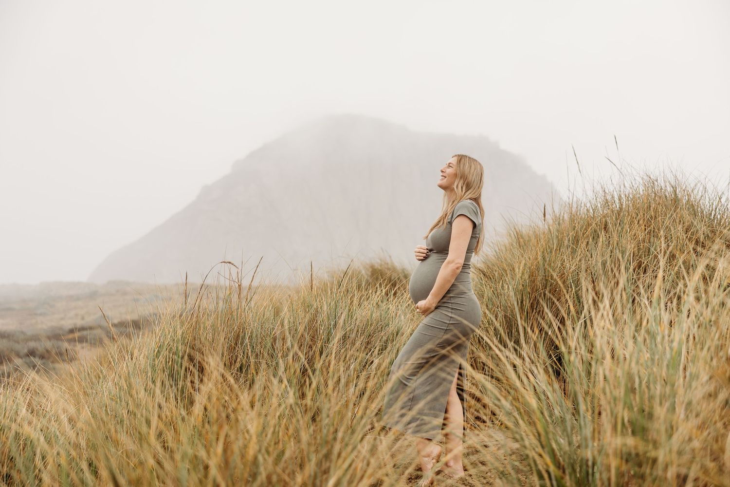 Maternity photo in warm tones featuring tall grass and mountains in the background.