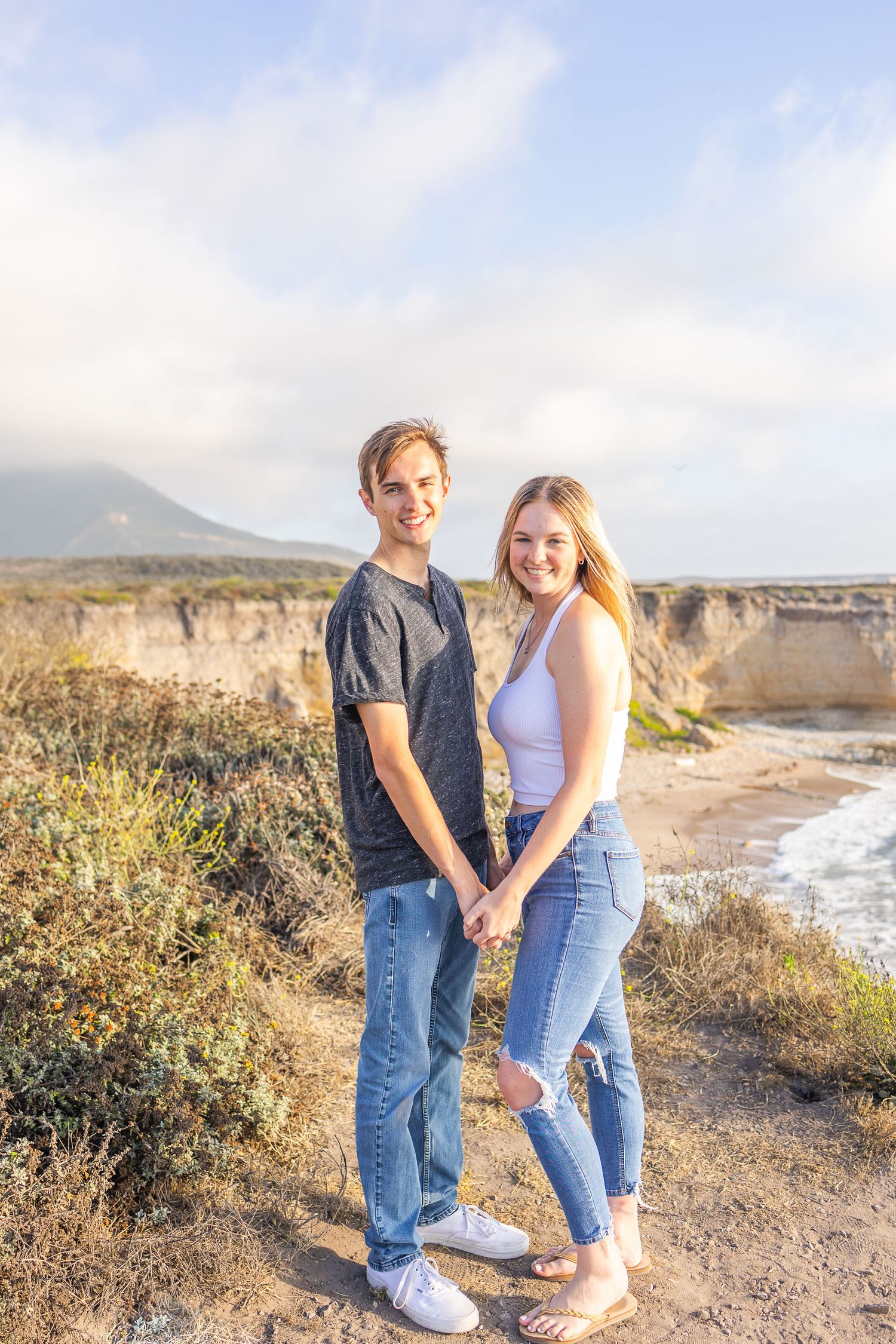 A couple holds hands while standing on a coastal cliff with ocean views behind them.