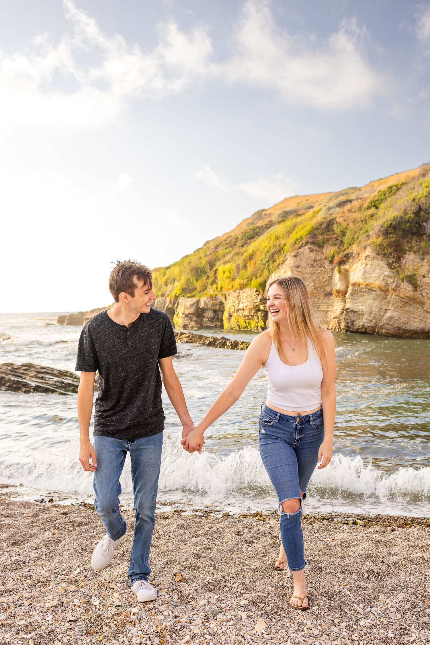 A couple walks hand in hand along a scenic beach with golden cliffs in the background.