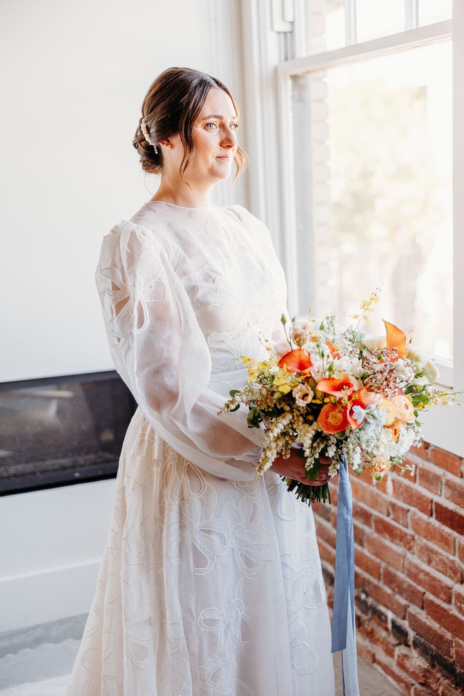 Bride in vintage-inspired gown with balloon sleeves holds coral and peach bouquet by window of Hotel Cerro San Luis Obispo.