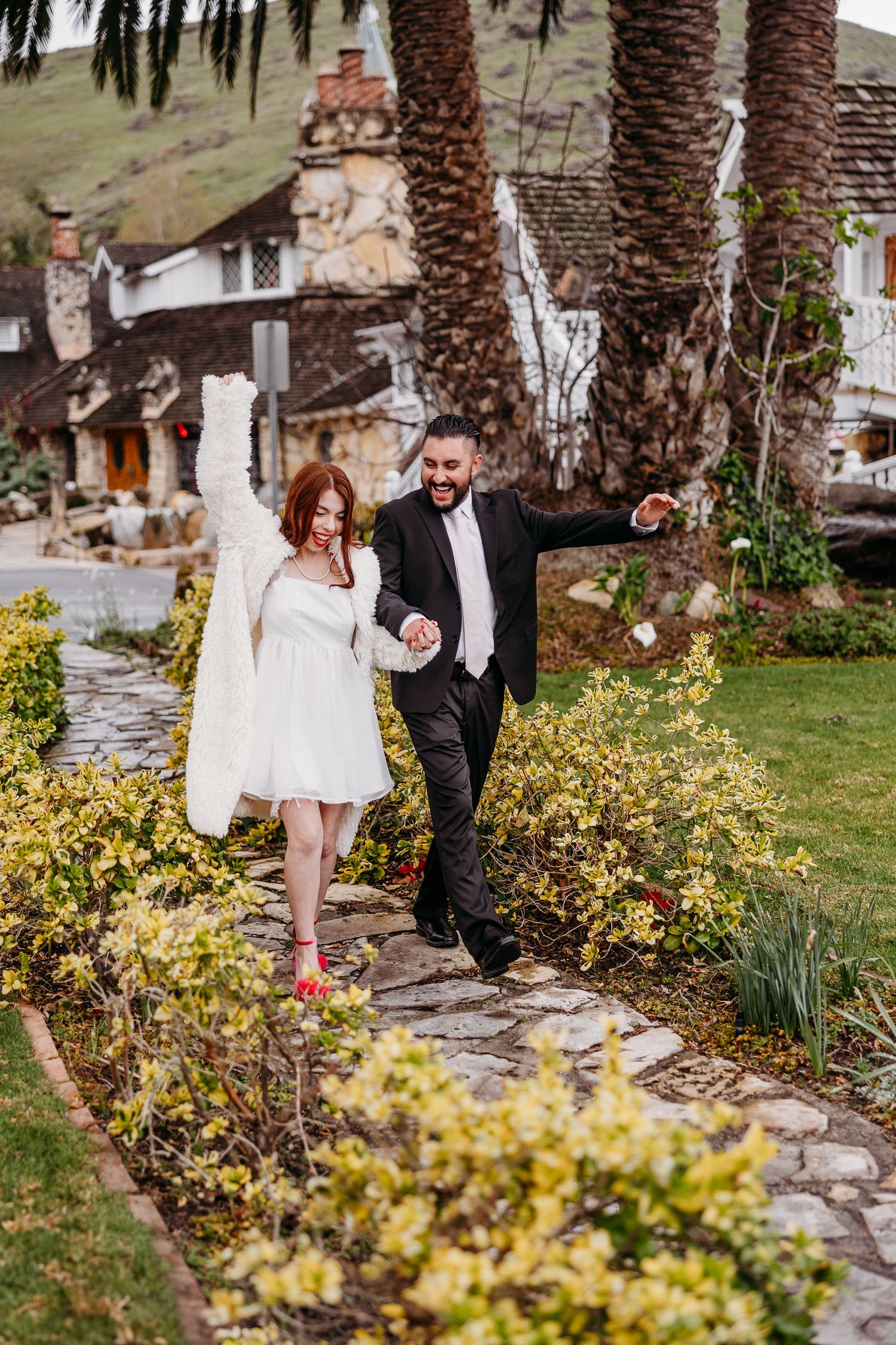 A couple walking along a stone path in a garden with palm trees and yellow flowers beside a rustic building.