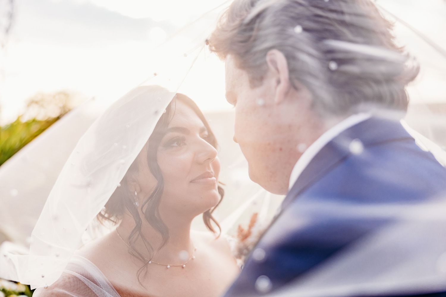 Romantic sunlit moment between newlyweds under bridal veil.