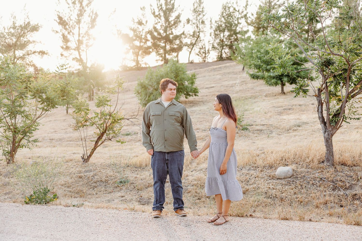 A couple strolls together on a sunny path at sunset in a rustic outdoor setting with scattered trees and rocks.