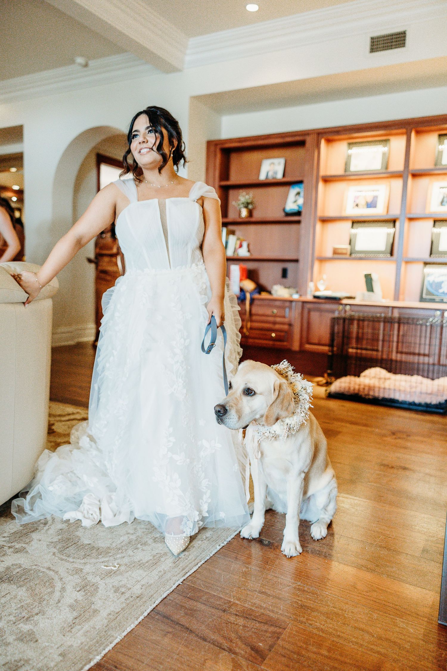 A bride in a white wedding gown stands with her yellow Labrador dog wearing formal attire in an elegant indoor setting.