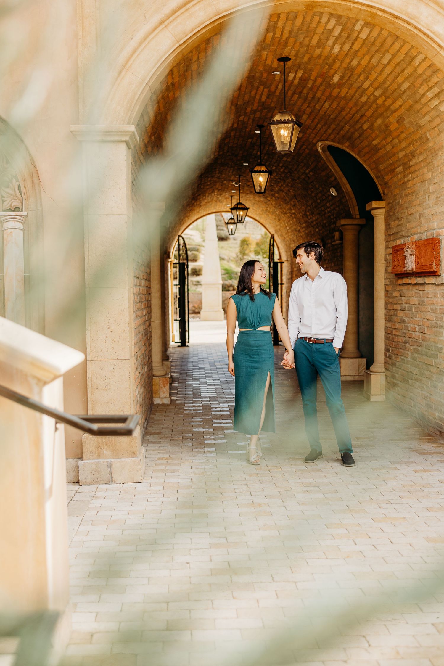 Romantic couple walking through a brick archway corridor with vintage lanterns overhead.