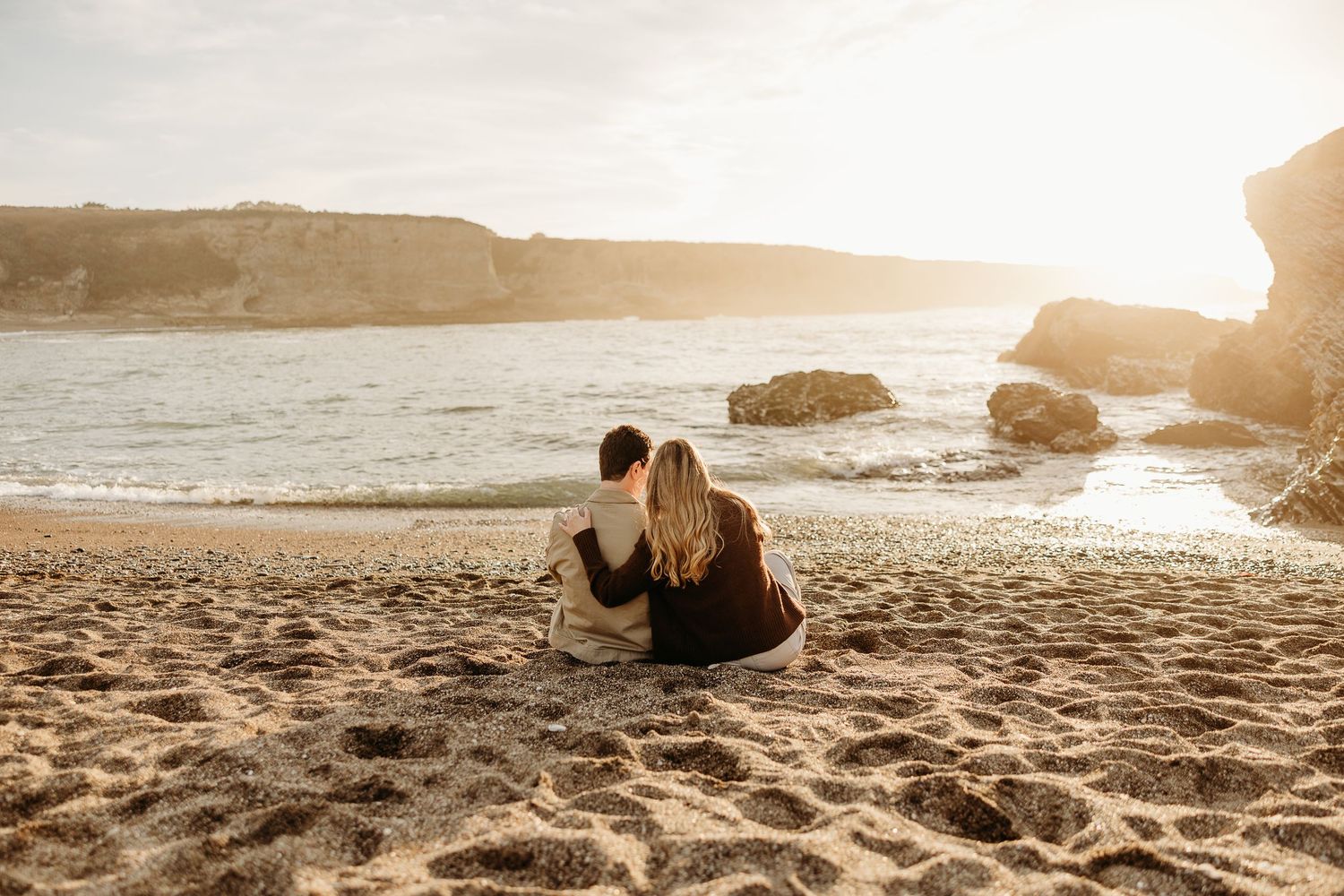 Wide panoramic shot of couple enjoying sunset on secluded beach with rocky outcrops.