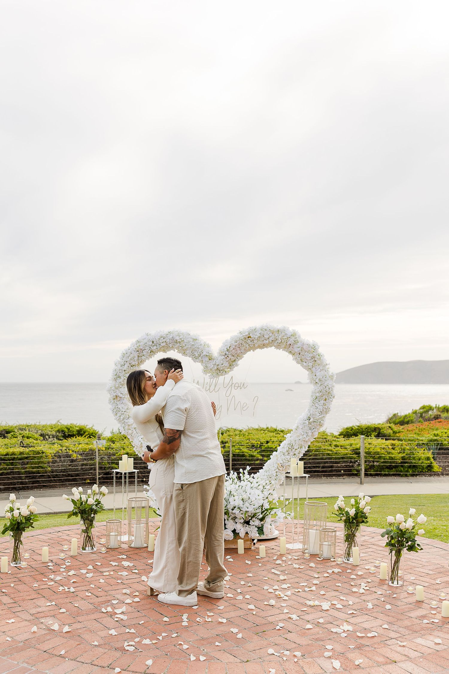 Outdoor wedding ceremony setup with white floral arch and rose petals scattered on brick patio overlooking scenic landscape.