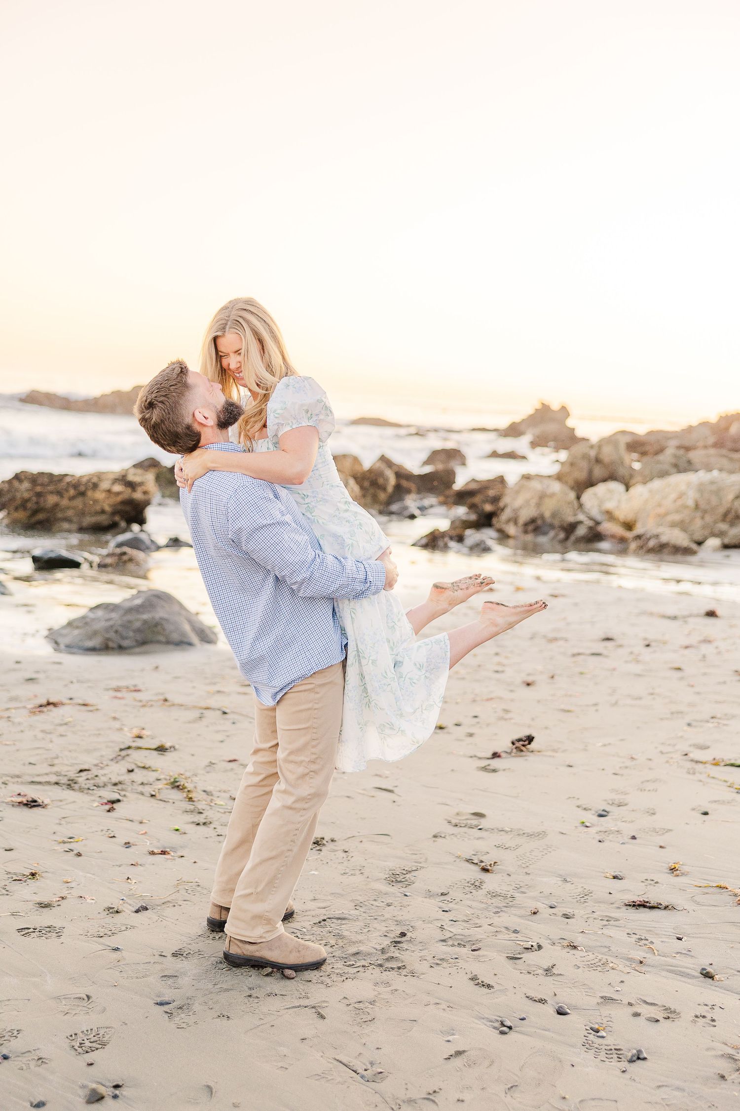 A playful couple dances together on a beach with rocky outcrops during sunset.