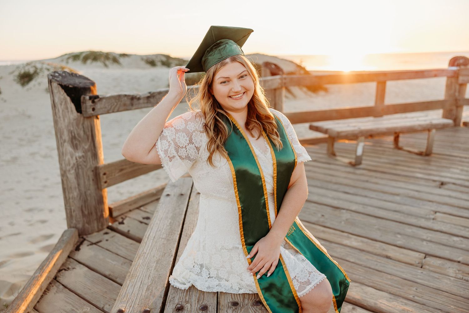 Graduate in cap and gown poses on wooden boardwalk during sunset beach photoshoot.