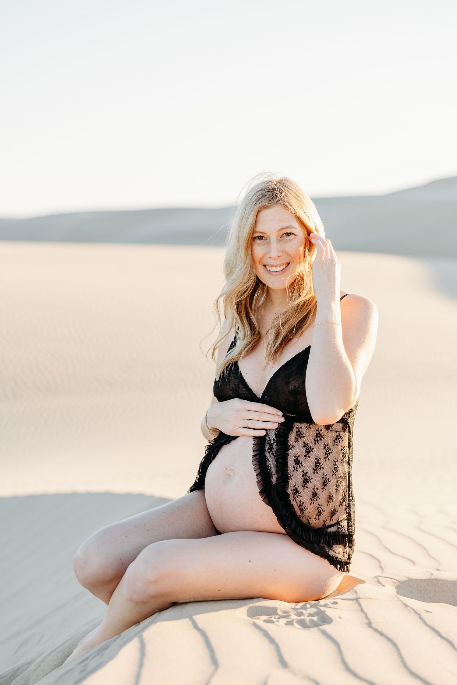 Portrait of person in black lace lingerie sitting on sand dunes at sunset.