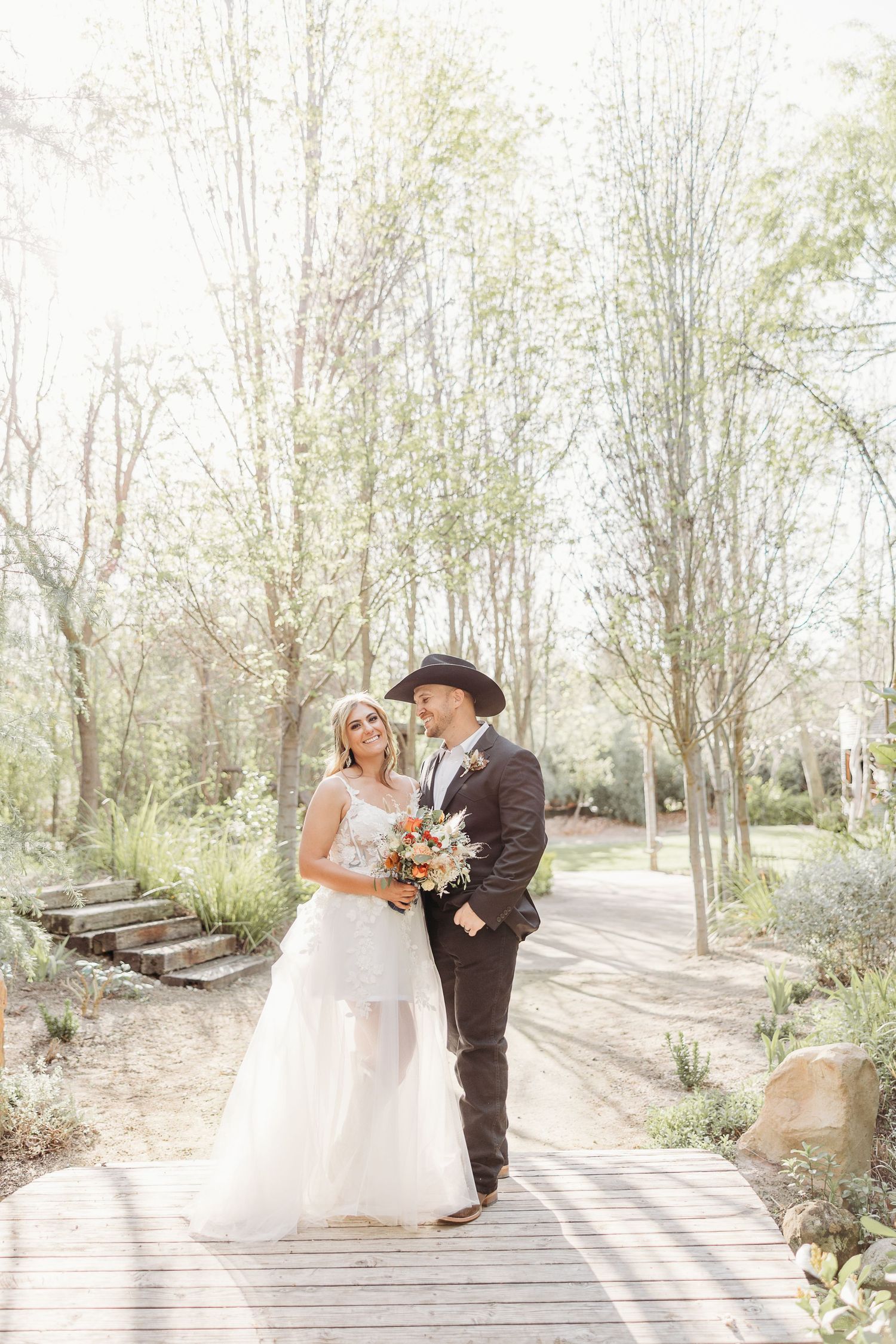 Bride and groom in western attire pose romantically on wooden pathway surrounded by trees.
