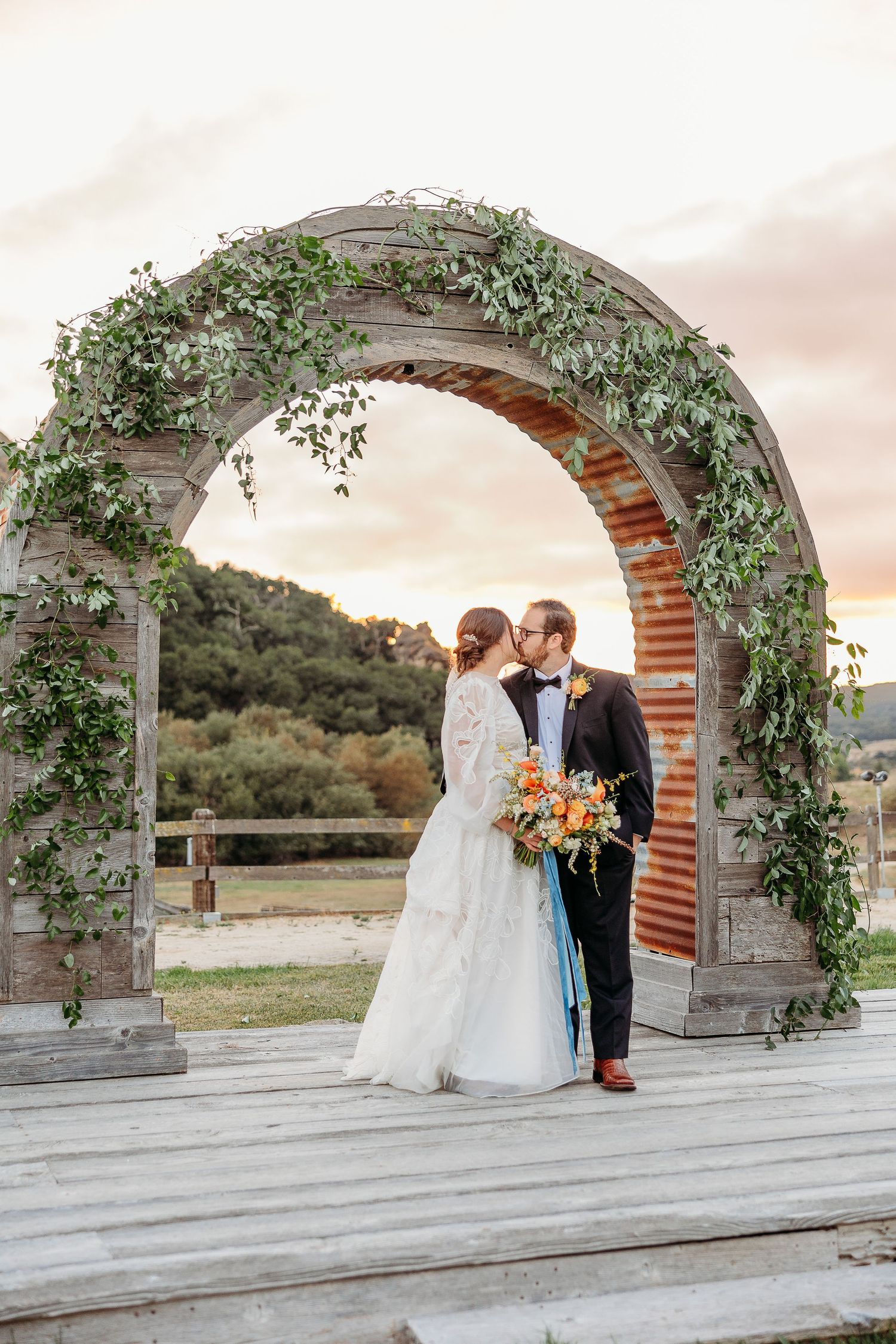 Wedding couple stands beneath an ivy-covered stone arch during golden hour celebration.