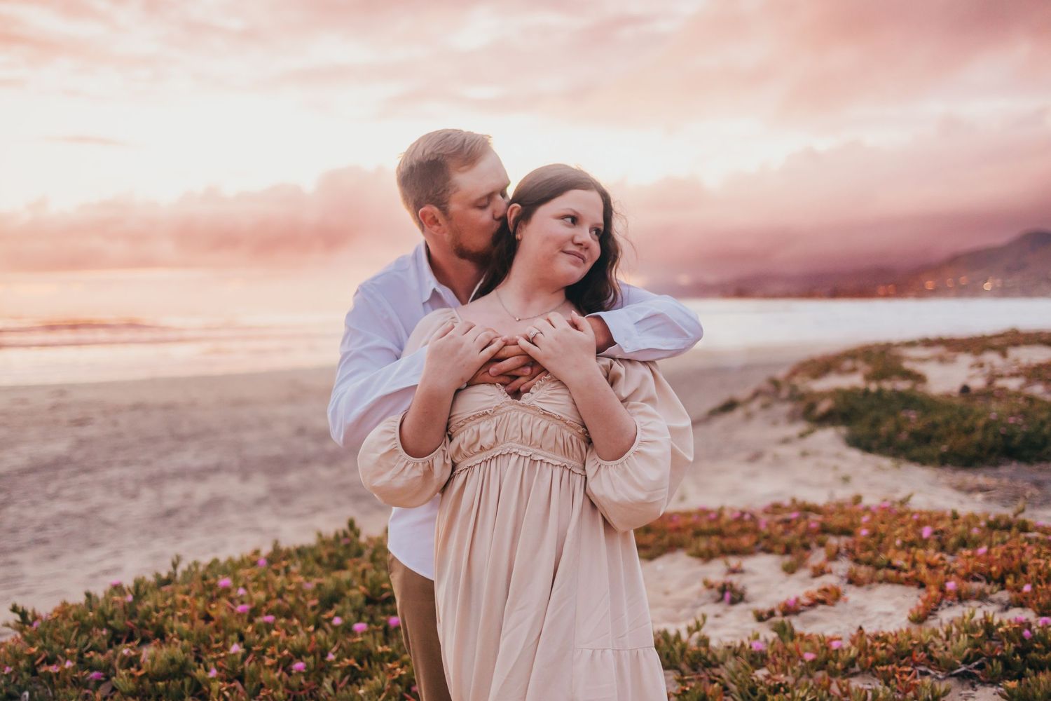 Couple embracing during golden hour on flower-dotted coastal cliffs.
