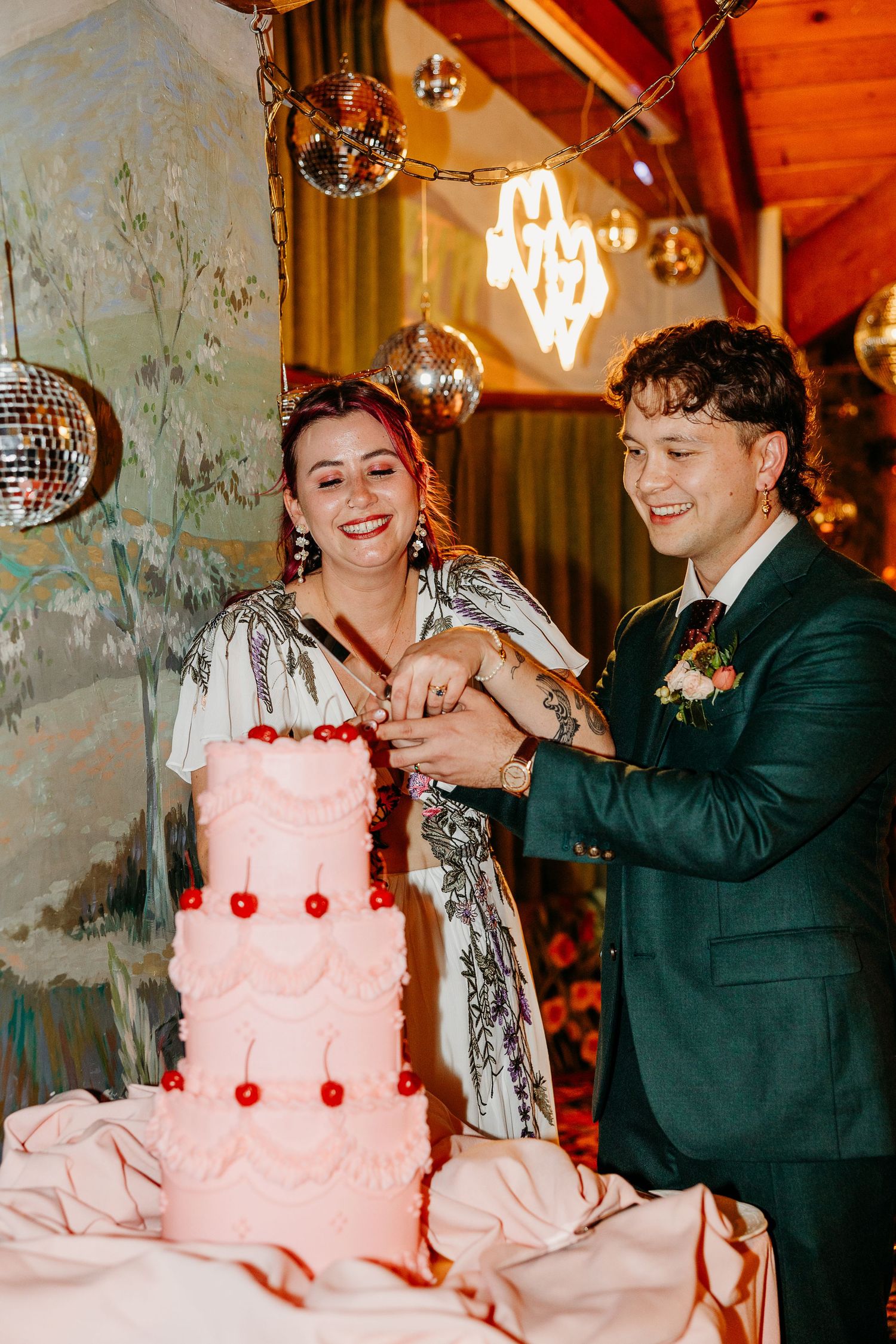 Couple cutting into a three-tiered pink wedding cake with cherry decorations under disco balls.