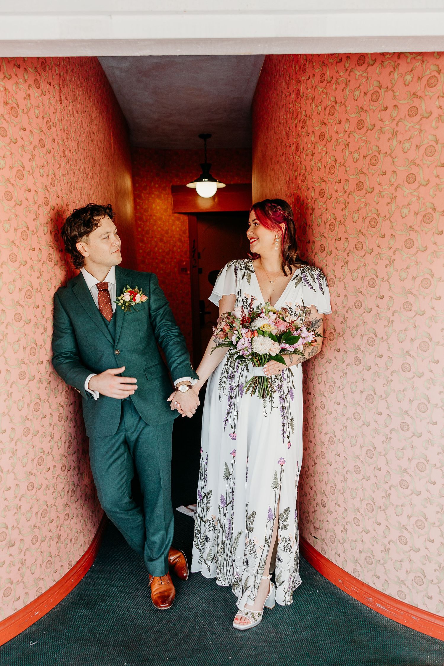 Wedding couple posing against coral wallpaper in a vintage-style hallway.