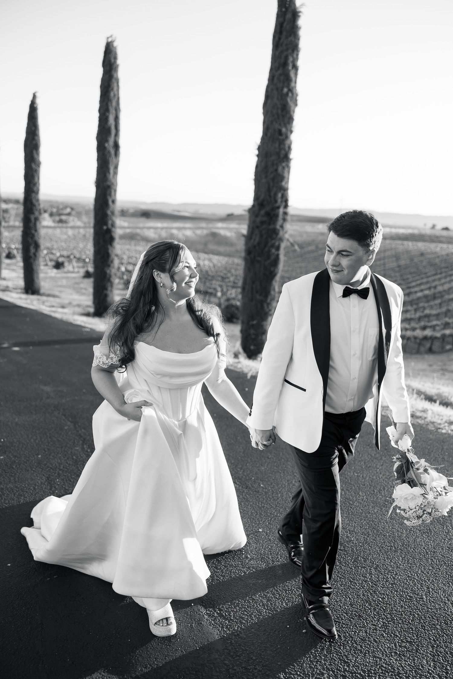 Black and white photo of bride and groom walking hand in hand along vineyard road.