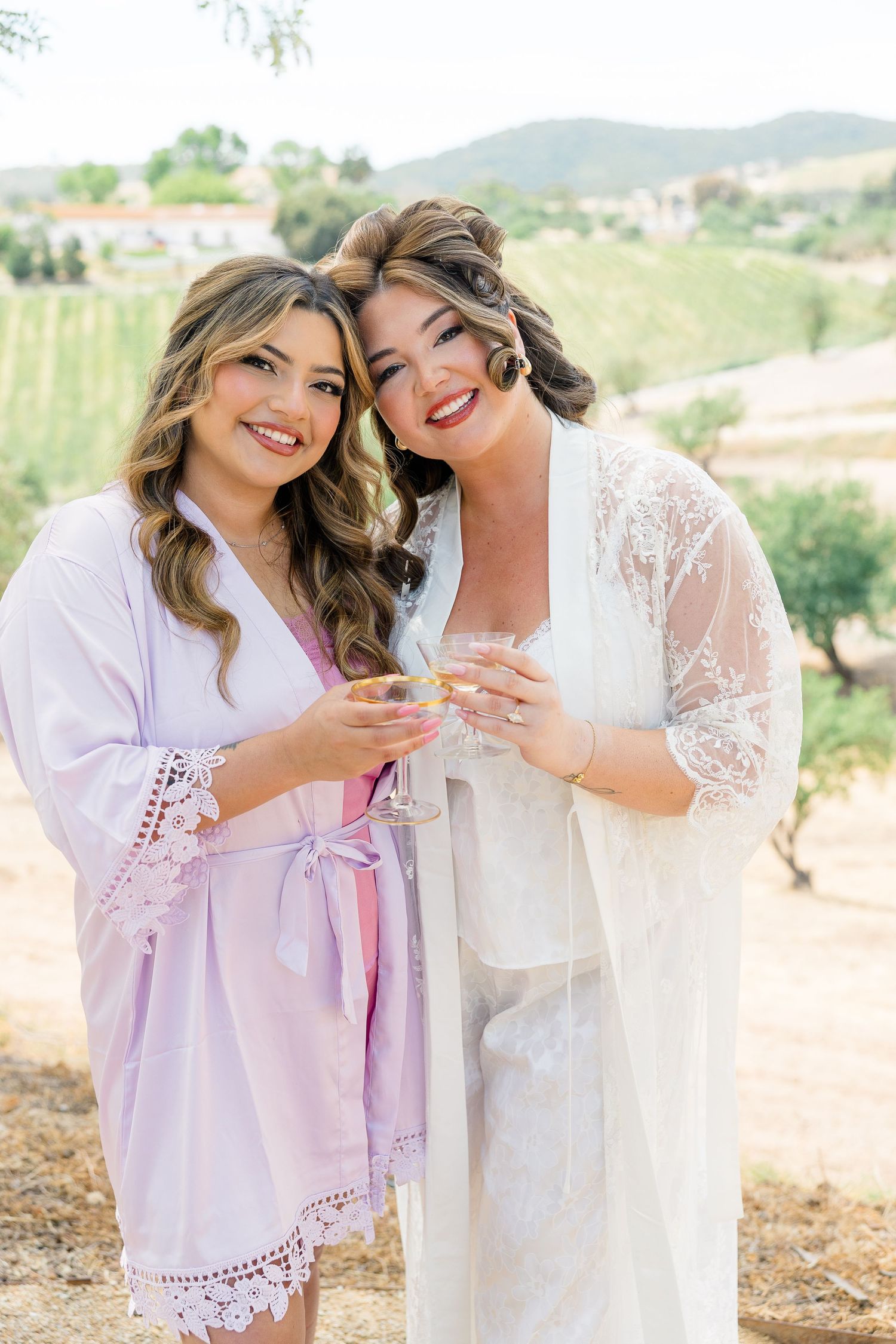 Two bridesmaids in lace robes toasting with champagne glasses at a vineyard wedding getting ready celebration.