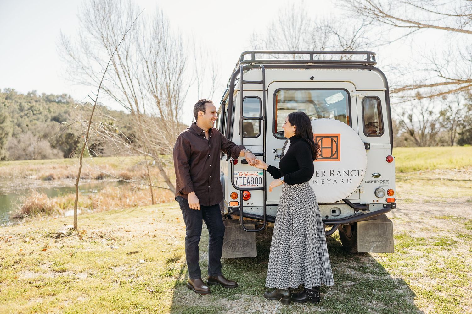 A couple poses together near a vintage Land Rover Defender in a scenic outdoor setting during a photoshoot.