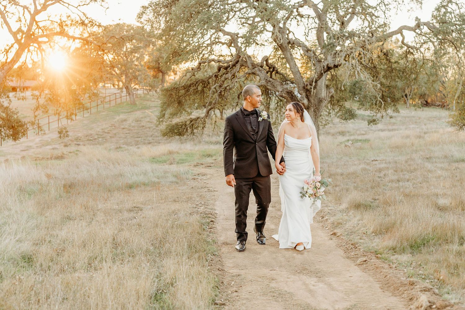 Wedding couple walking together at sunset through a golden field with trees in the background.