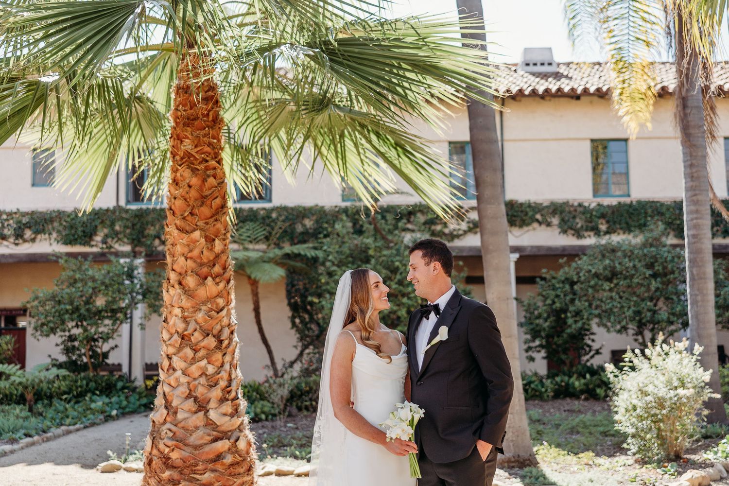 A romantic wedding portrait of newlyweds standing beneath palm trees in a Mediterranean-style courtyard.