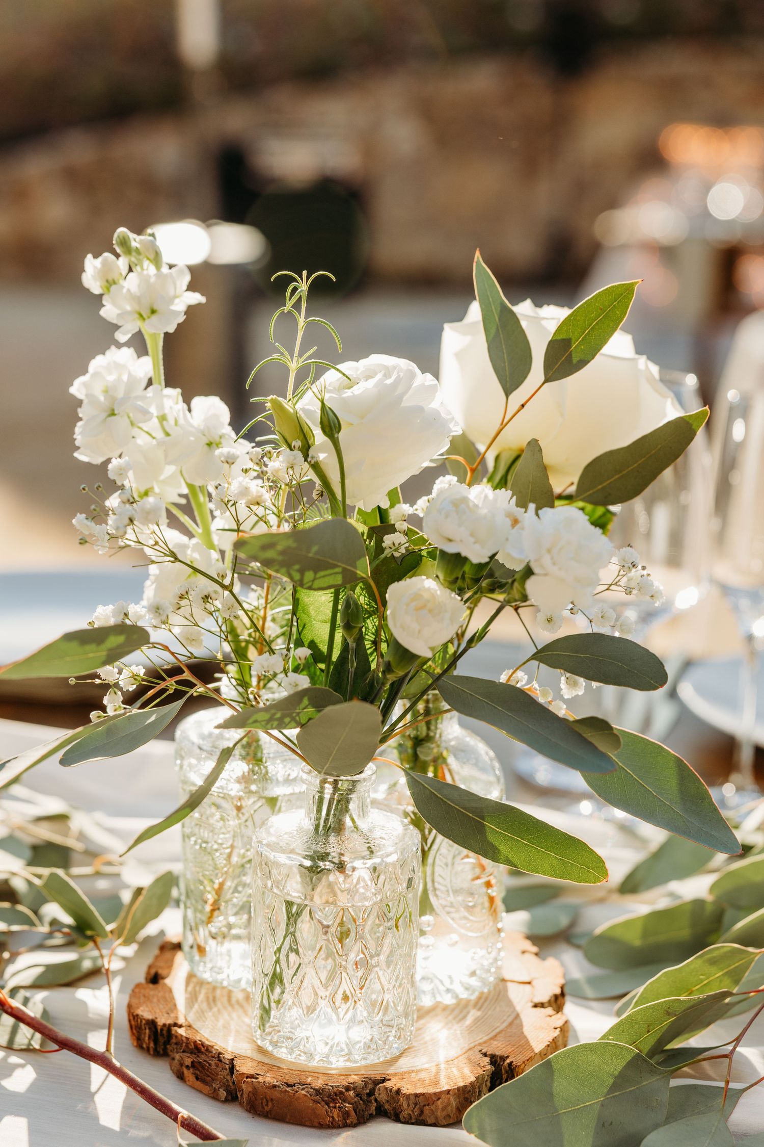 Elegant white floral centerpieces with greenery arranged on wood slices in glass jars.