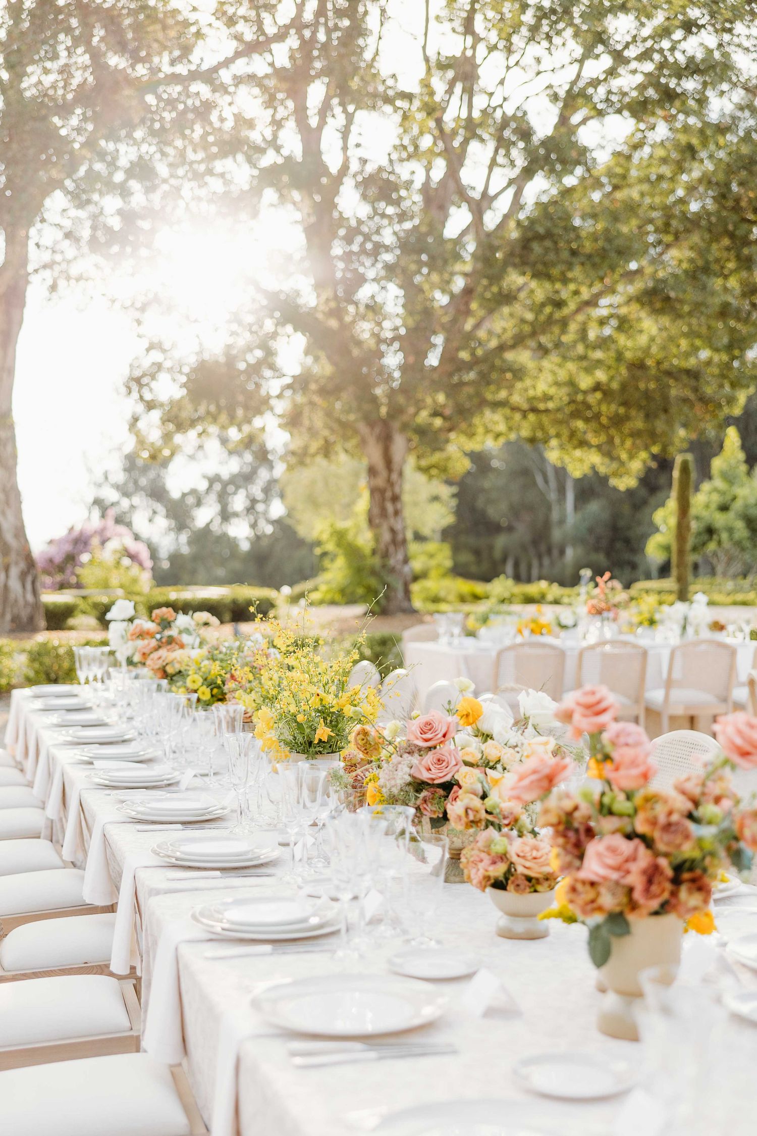 Elegant outdoor wedding reception table settings with pink floral centerpieces and white chairs under a canopy of trees.