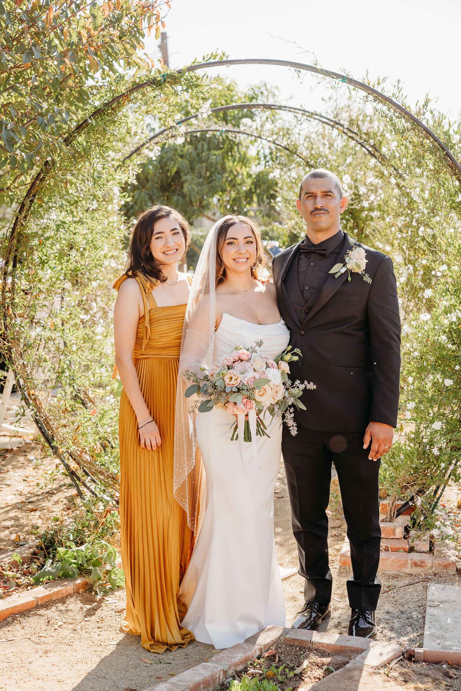 A wedding party poses under trees with a bridesmaid in a mustard yellow dress and a bride in white.